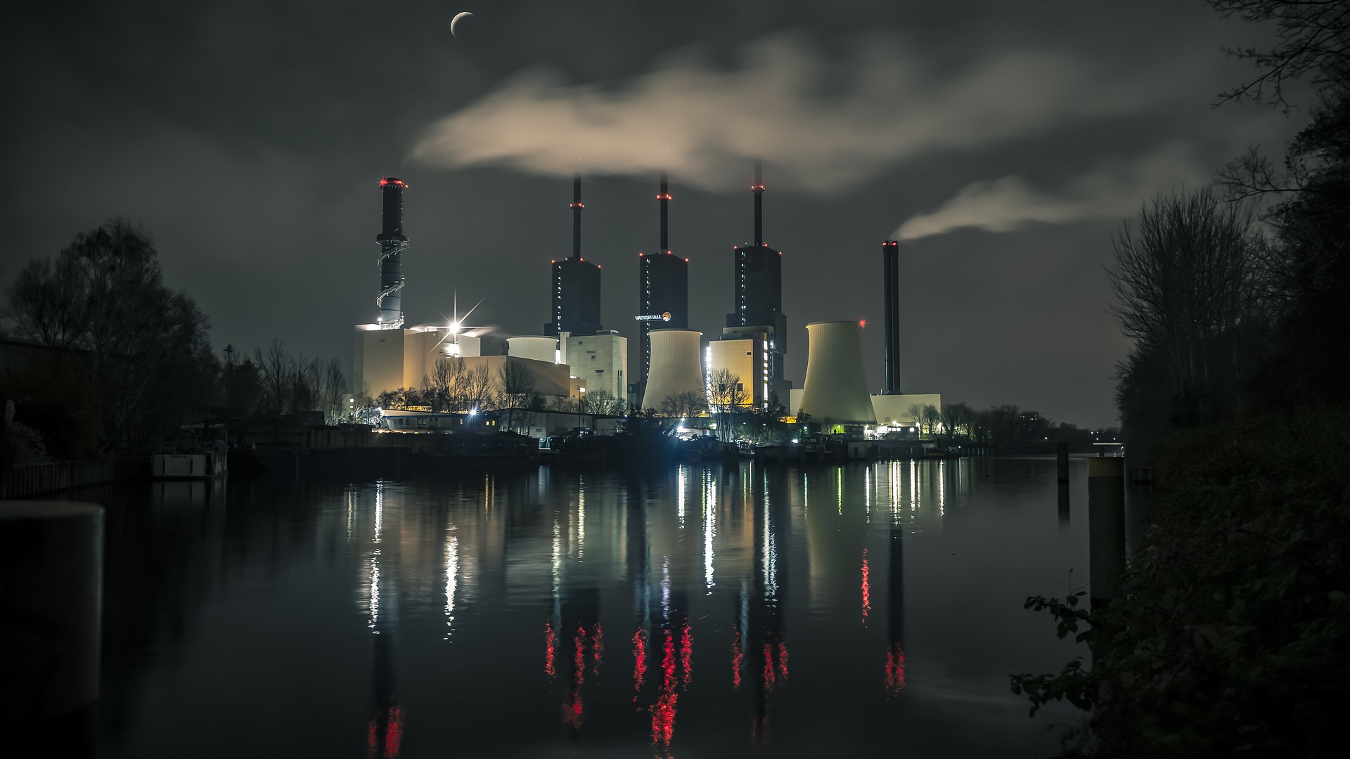 imagem de rio rodeado por mata durante a noite e, ao fundo, uma usina hidrelétrica iluminada com três torres e dois reatores.